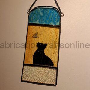 Stained glass wall hanging with three panels: a blue patterned top section, a golden amber middle panel featuring a black cat silhouette looking up at a small butterfly, and a textured cream base, suspended by a chain against a plain wall.