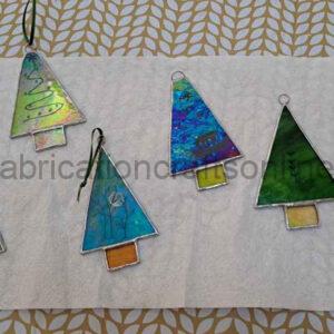 Five stained‑glass ornaments shaped like stylized Christmas trees rest on a white textured surface with a gold leaf‑patterned background. Each tree has a triangular body and a small rectangular base, featuring different colors and patterns: green and yellow swirls, blue abstract designs, solid green, turquoise with a leaf motif, and a blue‑purple blend. Small loops at the top allow them to be hung, with one ornament already attached to a black string.