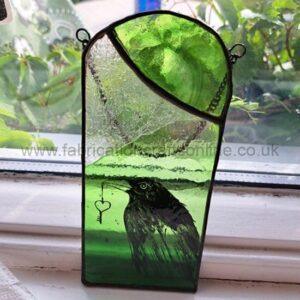 Green stained glass panel shaped like an arched window, featuring a black bird perched against layered green glass, with a delicate cobweb detail in the top corner, displayed on a windowsill with leafy plants outside.