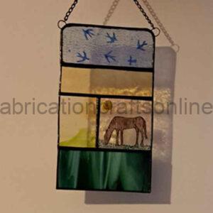 A stained‑glass panel hangs from a chain on a wall. The top section shows pale blue glass with dark blue silhouettes of birds in flight. The middle is split: the left side is a warm yellow panel, and the right side depicts a brown horse grazing on green grass beneath a yellow sun. The bottom section is a solid dark green rectangle. The colours and simple shapes create a calm pastoral scene.