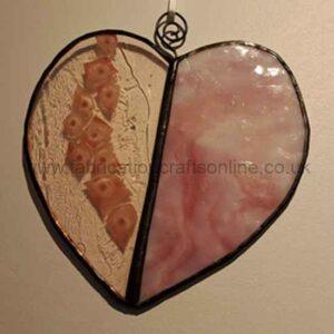 A heart‑shaped stained‑glass ornament divided vertically into two halves: the left side is textured beige with circular fossil‑like patterns, and the right side is smooth marbled pink with white swirls. A small wire loop with a spiral sits at the top for hanging, set against a light-colored wall.