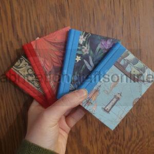 A hand holding four books all A7 in size. Two have red spines, two blue, and each has a different decorative paper on the cover