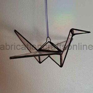 A minimalist metal sculpture shaped like an origami-style bird hangs from a thin string against a plain wall. The design is made from thin metal rods forming angular, geometric lines that outline the crane-like figure.