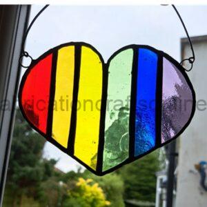 A heart‑shaped stained‑glass ornament hangs in front of a window. The heart is divided into vertical rainbow‑coloured segments—red, orange, yellow, green, blue, and purple—outlined in black solder. Soft daylight shines through the translucent glass, and trees and a building appear blurred in the background.