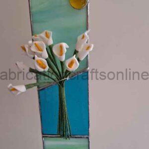 A vertical stained‑glass panel divided into three sections: pale green at the top and bottom and a vivid blue band across the middle. A small circular yellow glass piece sits near the top. At the center, a three‑dimensional bouquet of white calla lilies with yellow centers is mounted onto the glass, their green stems gathered and extending downward into the blue section. The flowers appear sculptural and lifelike, creating depth against the flat stained‑glass background.