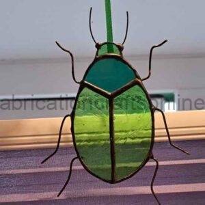 A stained‑glass ornament shaped like a beetle hangs from a green ribbon. Its body is made of green and teal glass segments outlined in metal, creating a layered, iridescent look. Thin wire forms the beetle’s legs and antennae, giving it a delicate, handcrafted feel. Light from a nearby window shines through the translucent glass, highlighting the colours and textures.