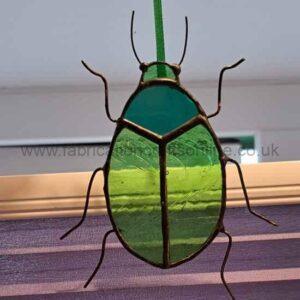 A stained‑glass ornament shaped like a beetle hangs from a green ribbon. Its body is made of green and teal glass segments outlined in metal, creating a layered, iridescent look. Thin wire forms the beetle’s legs and antennae, giving it a delicate, handcrafted feel. Light from a nearby window shines through the translucent glass, highlighting the colours and textures.