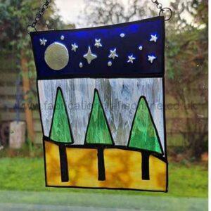 A stained‑glass panel hanging in a window, depicting a stylized landscape with three green triangular trees on yellow and brown ground beneath a blue sky. A silver moon and several white stars appear above the scene. Sunlight from outside illuminates the colors, with grass and a wooden fence visible through the window behind it.