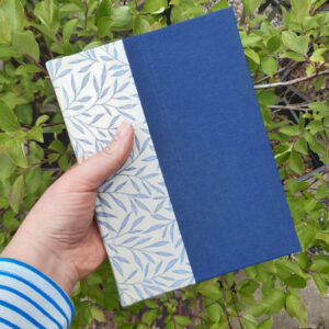 A hand holding a book with blue willow pattern spine and solid blue book cloth on the covers.
