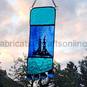 A rectangular stained‑glass hanging artwork with turquoise and deep blue panels, featuring a silhouetted pair of whimsical, pointed structures with swirling details. Five small blue glass beads dangle from the bottom edge. The piece hangs outdoors against a backdrop of trees and a cloudy sunset sky.