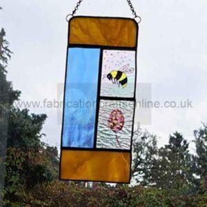 A stained‑glass panel hanging outdoors, divided into five sections with black borders. The top and bottom sections are amber glass, the left middle section is light blue, and the right middle is split into two smaller panels: one showing a yellow‑and‑black bee with translucent wings, and the other depicting a pink and yellow flower with a green stem on textured clear glass. Trees and a cloudy sky appear in the background.