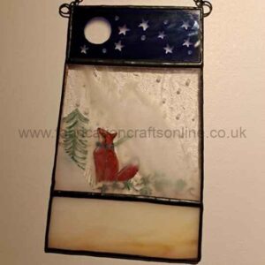 A stained‑glass panel divided into three sections: a dark blue night sky with a white moon and stars at the top, a red fox sitting in snow beside a green pine tree in the middle, and a warm-toned gradient at the bottom. The piece is framed in black with two loops for hanging.