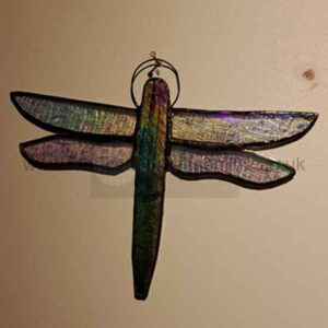 An iridescent dragonfly ornament with green, purple, and blue metallic tones hangs against a beige wall. It has two pairs of wings, a long narrow body, and a small wire loop at the top for mounting.