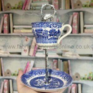 Blue-and-white porcelain teacup on a tall ornate stand with a matching saucer, held against a blurred bookshelf in the background.