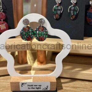 Colorful mosaic-style round drop earrings with silver studs hanging from a white display frame on a wooden stand; background shows more earrings and a label reading 'Hold me up to the light'.