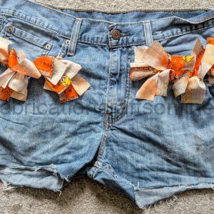 Blue denim shorts decorated with orange and beige fabric ribbons along the waistband, laid on a carpeted floor.