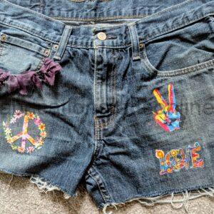 Blue denim shorts with a frayed hem, purple fabric ruffle near the left pocket, and bright embroidered patches on the legs (flower wreath on the left, colorful peace-hand on the right).