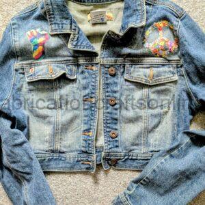 Blue denim jacket laid on a beige carpet with two patches: a multicolored raised fist on the left chest and a floral peace sign on the right chest.