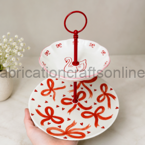 Two-tier white ceramic serving stand with red ribbon and heart designs, held by a hand over a light countertop.