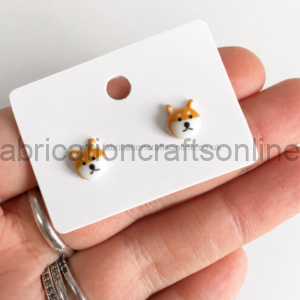 Tiny Shiba Inu face stud earrings on a white display card held in a hand.