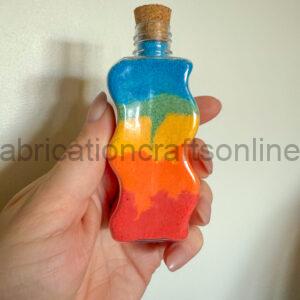 Small glass bottle with a cork stopper, filled with layered rainbow-colored sand, held in a hand against a plain wall.