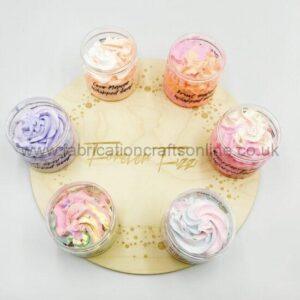 Six small glass jars of pastel whipped creams arranged in a circle on a light wooden board, labeled flavors like coconut mango and fruit swirl.