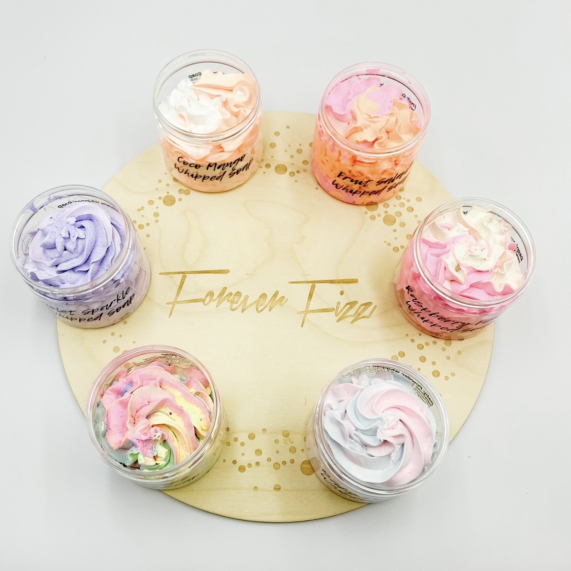 Six small glass jars of pastel whipped creams arranged in a circle on a light wooden board, labeled flavors like coconut mango and fruit swirl.