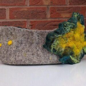 Grey felted headband featuring a large multicolored flower (yellow, green, blue) on the side, displayed on a white surface against a brick wall.