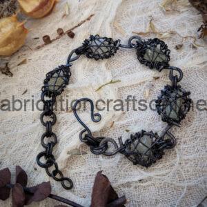 Handmade black wire-wrapped bracelet with five green stones and an S-hook clasp, laid on beige fabric.