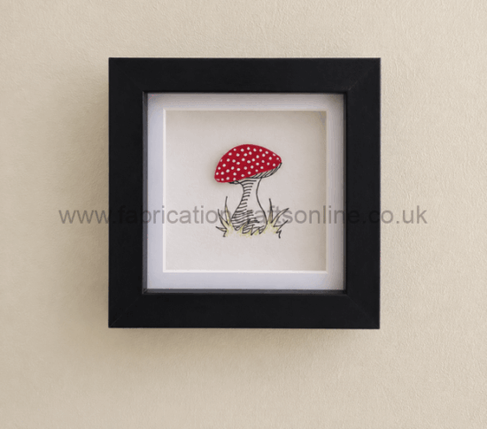 Framed artwork of a red mushroom with white spots on a beige wall, black frame, small sketchy grass at base.