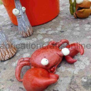 Red clay crab sculpture with white eyes sits on a weathered pavement, surrounded by tall gray tree-like figures and small orange creatures in an outdoor craft display.