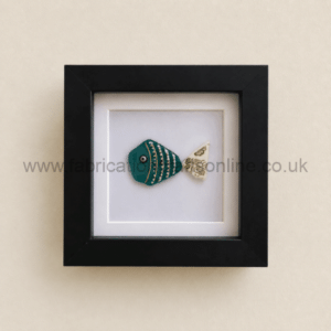 Framed ceramic teal fish with gold stripes and a patterned white tail, mounted in a black square frame on a beige wall.