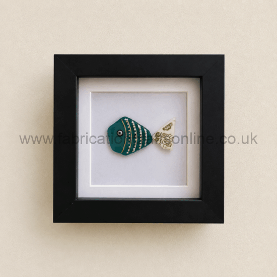 Framed ceramic teal fish with gold stripes and a patterned white tail, mounted in a black square frame on a beige wall.