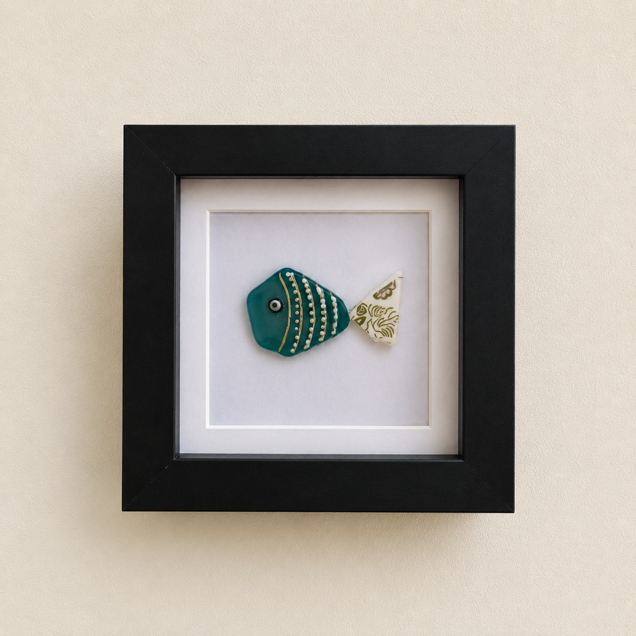 Framed ceramic teal fish with gold stripes and a patterned white tail, mounted in a black square frame on a beige wall.