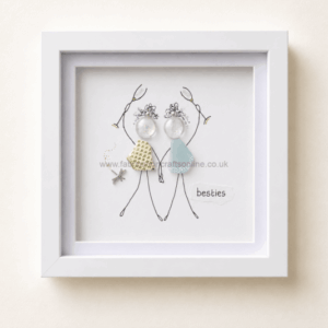 Framed illustration of two stick-figure friends holding hands, wearing fabric dresses, with decorative earrings and a star charm, captioned “besties”