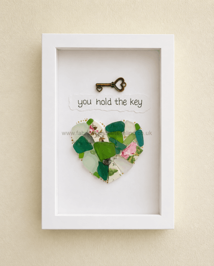 Framed art: a bronze heart-shaped key above a torn-paper caption 'you hold the key' with a mosaic heart of glass pieces.