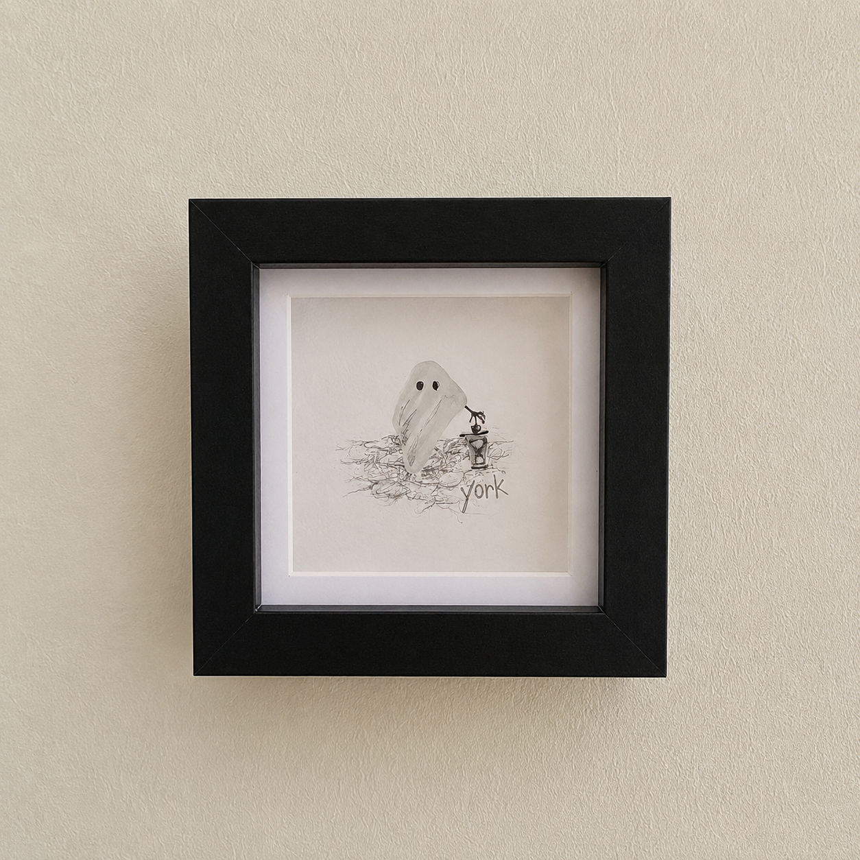 Small framed sketch of a ghost next to a person on a scribbled ground, inside a black square frame on a beige wall.