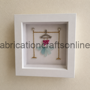 White display frame housing a miniature dress crafted from pink and pale blue glass pieces on a decorative metal hanger with gold chains outside the frame edge, like a jewelry display.