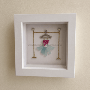 White display frame housing a miniature dress crafted from pink and pale blue glass pieces on a decorative metal hanger with gold chains outside the frame edge, like a jewelry display.