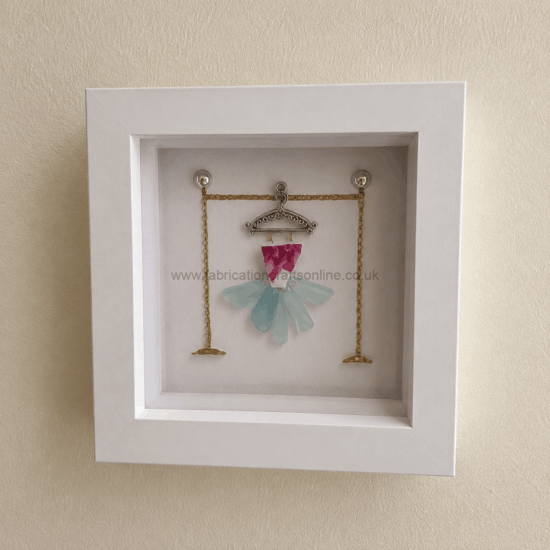 White display frame housing a miniature dress crafted from pink and pale blue glass pieces on a decorative metal hanger with gold chains outside the frame edge, like a jewelry display.