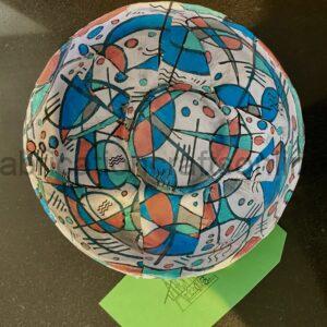 Colorful abstract painted ball with blue, teal, orange shapes and black lines on a dark surface, with a green sheet beneath.