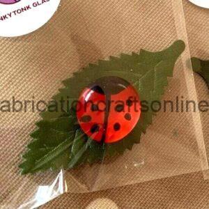 Red ladybug embellishment resting on a green leaf, inside a clear plastic bag on a beige textured surface.