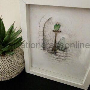 White shadow-box style frame holding textured artwork of a small green heart on a stand, with a plant visible to the left.