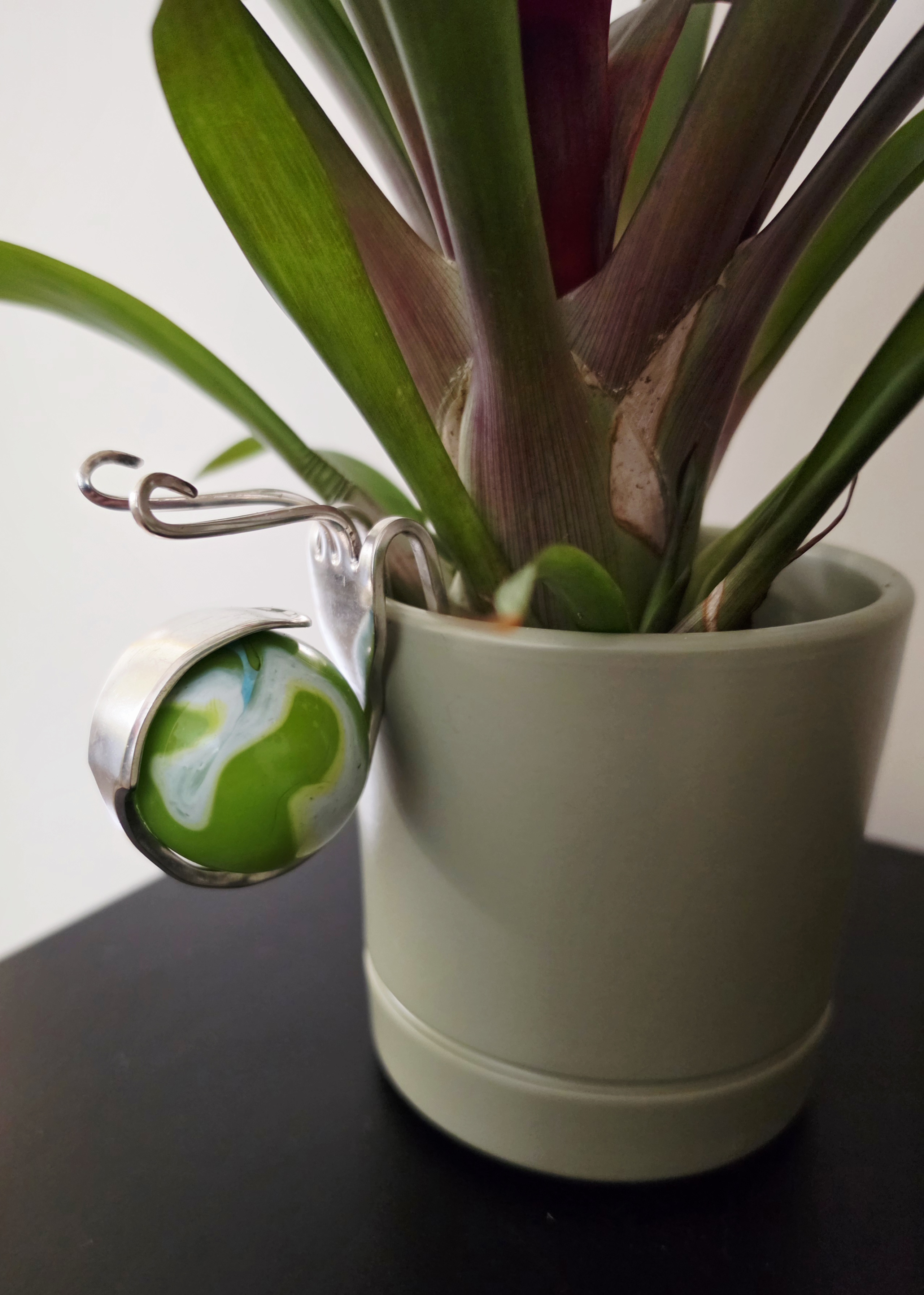 Close-up of a green and purple-stemmed plant in a gray ceramic pot, with a shiny silver hanging ornament featuring green and white swirls attached to the pot.
