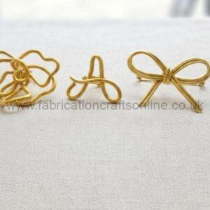 Three gold wire craft shapes on pale fabric: a tangled flower-like loop, the letter A, and a bow, arranged in a row.