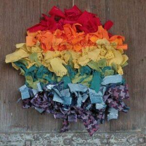 Rainbow-colored fabric strips arranged in horizontal bands on a wooden floor, forming a decorative patchwork mat or banner Readable colors from red to purple.