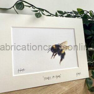 White picture frame with a watercolor bee illustration mounted on a white mat, resting on a wooden surface with trailing green plants nearby; captioned 'Honey, I'm Home' along the bottom edge.