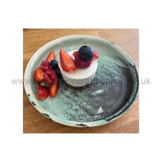 Small white cheesecake topped with blueberries, raspberries, and strawberry slices on a turquoise ceramic plate.