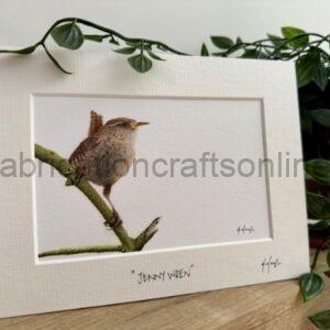 Framed watercolor of a small brown bird perched on a mossy branch, mounted on a white mat and leaning on a wooden surface with green plants nearby.