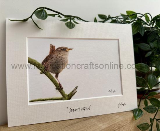 Framed watercolor of a small brown bird perched on a mossy branch, mounted on a white mat and leaning on a wooden surface with green plants nearby.